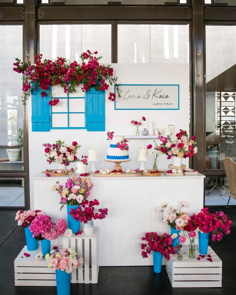 Floral first birthday dessert table with blue shutters, white cakes, and pink bougainvillea