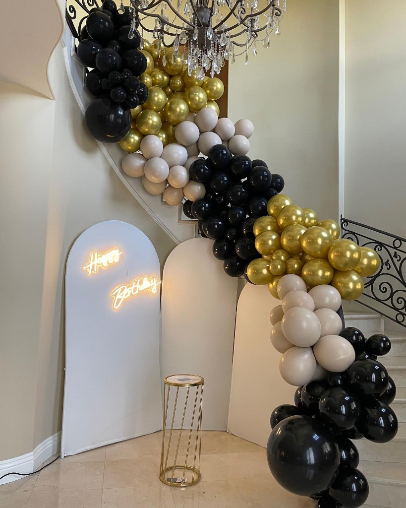 Black, gold, and beige balloon garland cascading down a staircase with Happy Birthday neon