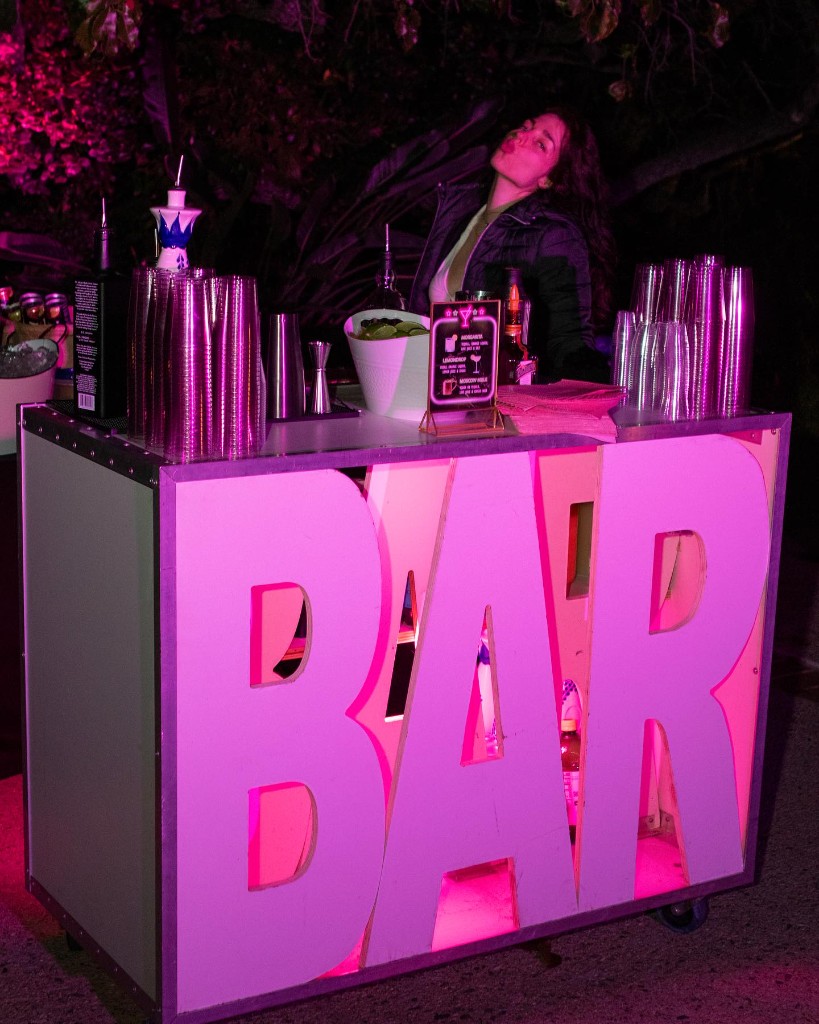 Bartender at a mobile bar with glowing pink BAR letters and cocktail menu at night