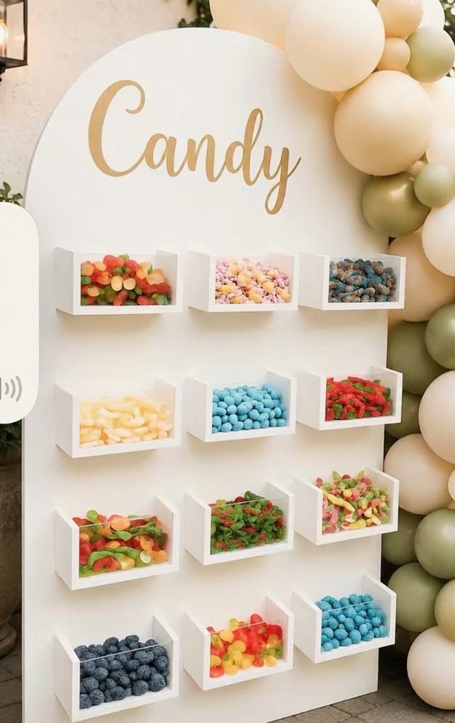 White arched candy wall with gold Candy lettering, twelve candy bins, and a cream and sage balloon arch