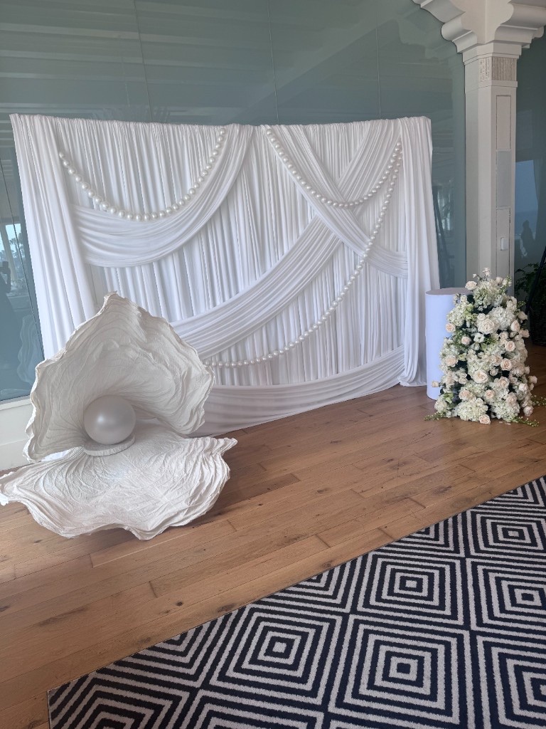 Pearl-draped white backdrop with giant clam shell prop and white florals